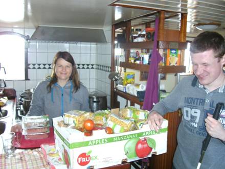 Foto: Katrin und Norbert in der Küche. Auf der Theke steht eine große Kiste mit allen Lebensmitteln für das Mittagessen (23 Personen). Beie lächeln. Liegt es vielleicht daran, weil die Kiste so groß ist, oder weil sie sich schon auf das essen freuen "g"?